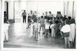 Uma fotografia em preto e branco que mostra uma fila de crianças em um ambiente interno ou coberto, possivelmente em uma escola ou centro comunitário. É possível identificar a presença de uma figura vestida de Papai Noel, sugerindo um evento de celebração natalina, comum em pequenas cidades brasileiras durante meados do século XX.