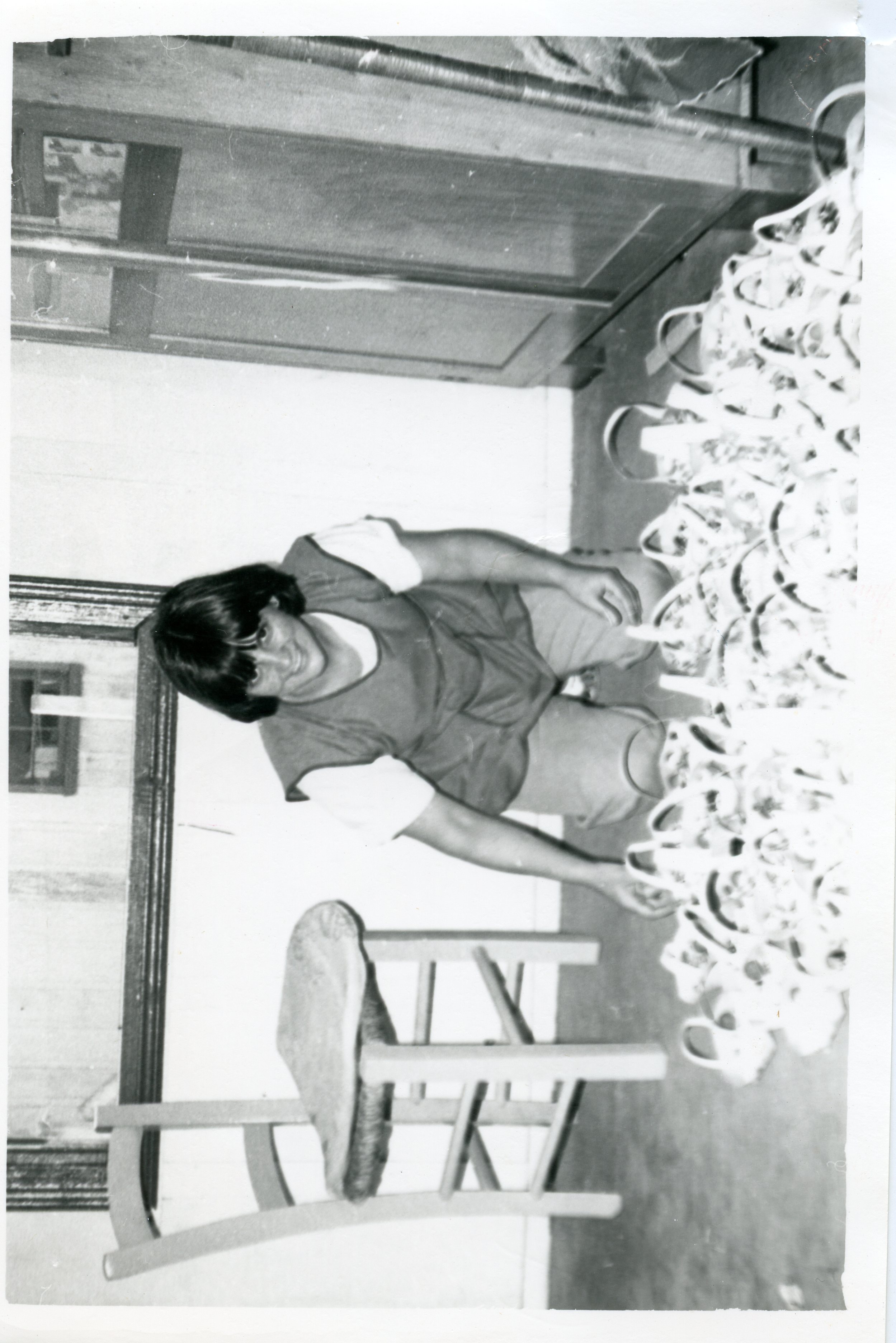 Uma fotografia em preto e branco que mostra uma mulher jovem agachada em um ambiente interno, possivelmente uma residência ou oficina, organizando uma grande quantidade de objetos circulares, como cestas ou moldes. A cena sugere uma atividade artesanal ou produtiva comum em pequenas cidades brasileiras, com a mulher vestindo roupas simples e casuais, indicando um período provável entre as décadas de 1970 e 1980.