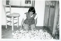 Uma fotografia em preto e branco que mostra uma mulher jovem agachada em um ambiente interno, possivelmente uma residência ou oficina, organizando uma grande quantidade de objetos circulares, como cestas ou moldes. A cena sugere uma atividade artesanal ou produtiva comum em pequenas cidades brasileiras, com a mulher vestindo roupas simples e casuais, indicando um período provável entre as décadas de 1970 e 1980.
