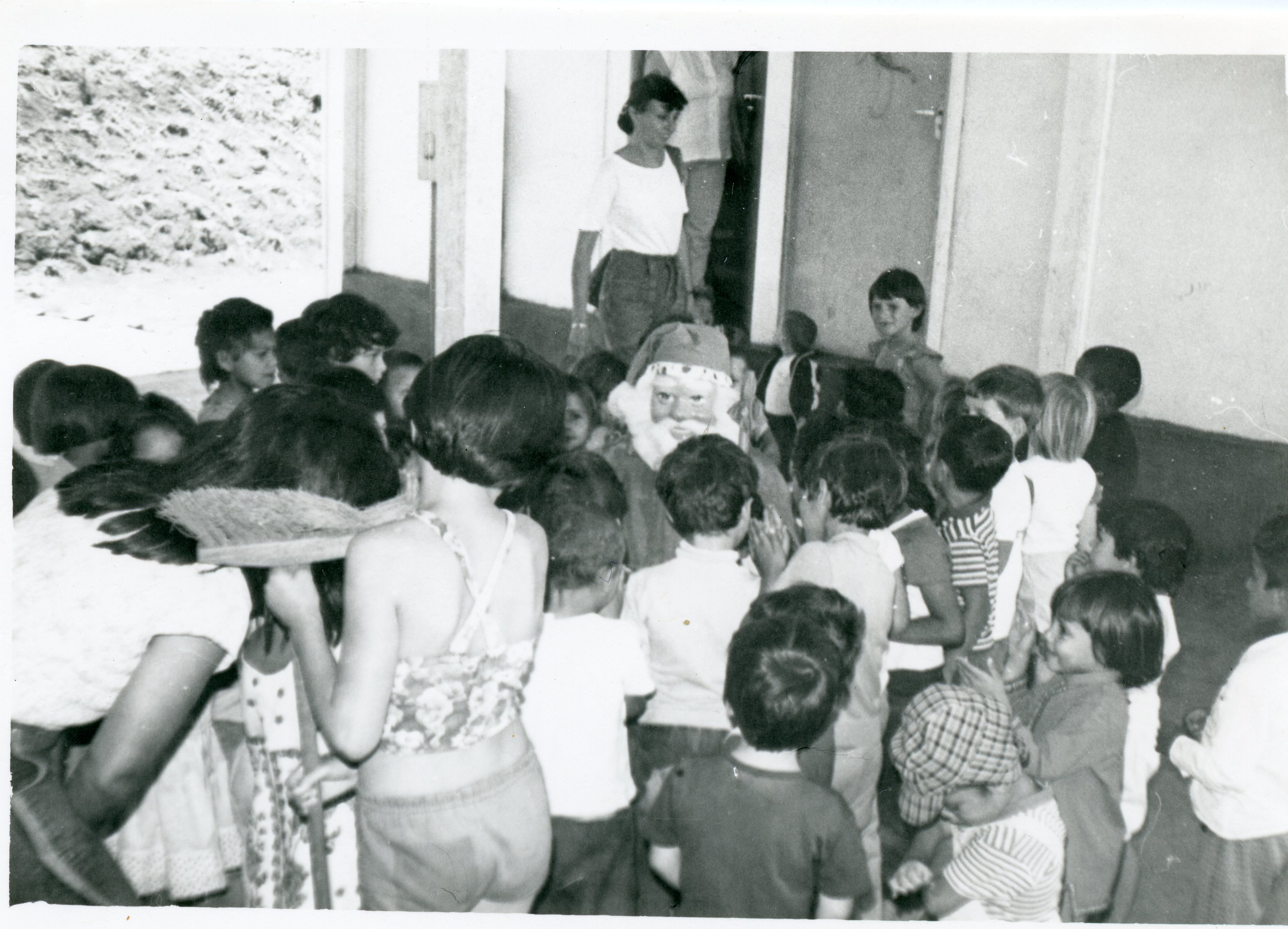 Uma fotografia em preto e branco que retrata uma celebração natalina em um ambiente escolar ou comunitário, com uma pessoa fantasiada de Papai Noel cercada por um grupo de crianças. A cena sugere uma atividade festiva típica de meados da década de 1980, refletindo o cotidiano de uma comunidade brasileira.