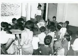 Uma fotografia em preto e branco que retrata uma celebração natalina em um ambiente escolar ou comunitário, com uma pessoa fantasiada de Papai Noel cercada por um grupo de crianças. A cena sugere uma atividade festiva típica de meados da década de 1980, refletindo o cotidiano de uma comunidade brasileira.