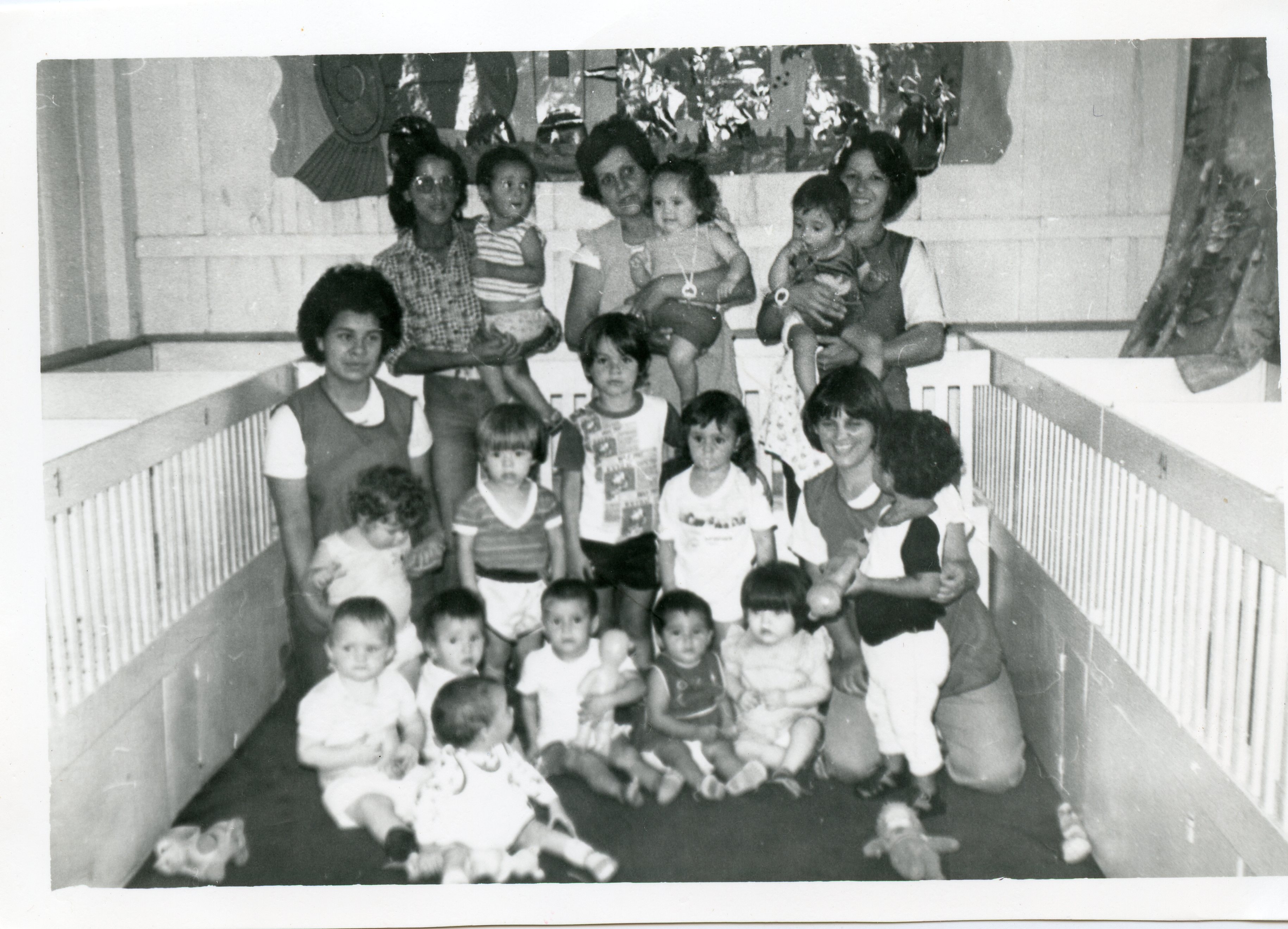 Fotografia em preto e branco retratando um grupo de mulheres e crianças em um ambiente que parece ser uma creche ou berçário, com berços de madeira visíveis nas laterais. A cena sugere uma atividade educacional ou de cuidado infantil, possivelmente em uma instituição brasileira entre as décadas de 1970 e 1980.