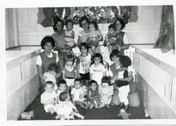 Fotografia em preto e branco retratando um grupo de mulheres e crianças em um ambiente que parece ser uma creche ou berçário, com berços de madeira visíveis nas laterais. A cena sugere uma atividade educacional ou de cuidado infantil, possivelmente em uma instituição brasileira entre as décadas de 1970 e 1980.