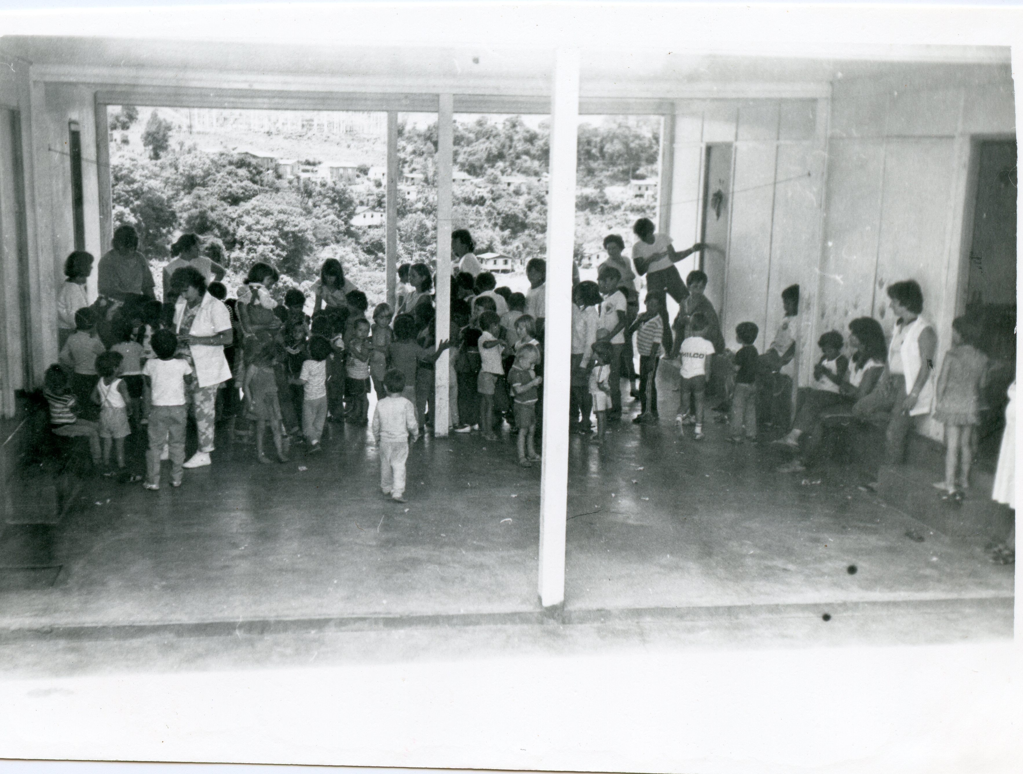 Fotografia em preto e branco registrando um grupo de crianças e adultos em um espaço semiaberto, possivelmente uma escola ou centro comunitário. O ambiente possui uma grande abertura lateral com vista para uma encosta arborizada, sugerindo uma região de relevo acentuado como o interior de Santa Catarina, possivelmente na década de 1970 ou 1980.
