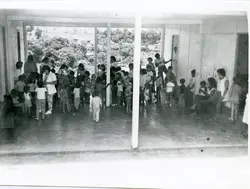 Fotografia em preto e branco registrando um grupo de crianças e adultos em um espaço semiaberto, possivelmente uma escola ou centro comunitário. O ambiente possui uma grande abertura lateral com vista para uma encosta arborizada, sugerindo uma região de relevo acentuado como o interior de Santa Catarina, possivelmente na década de 1970 ou 1980.