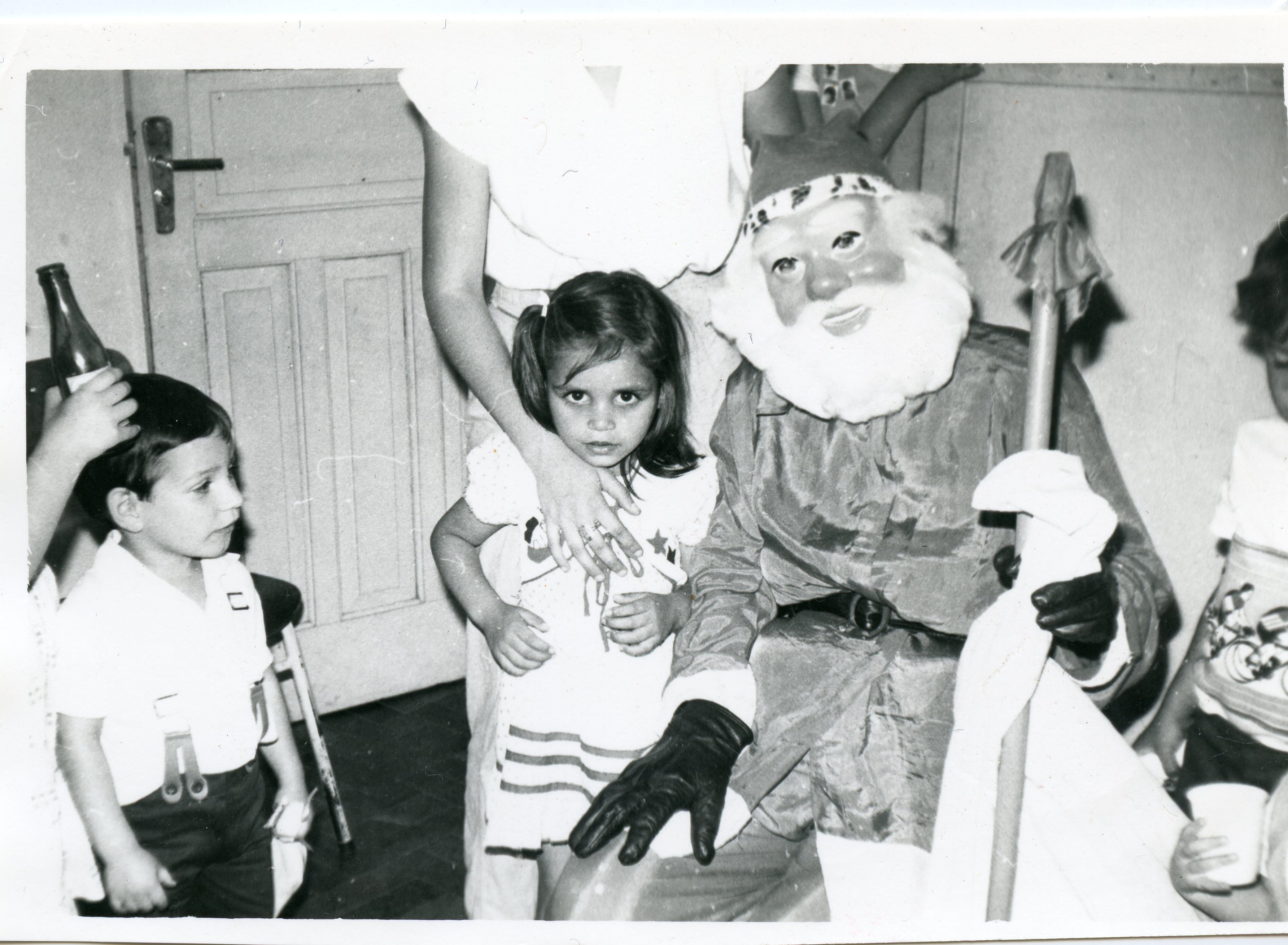 Fotografia em preto e branco capturando uma celebração natalina com uma criança posando ao lado de uma pessoa fantasiada de Papai Noel. O cenário sugere um ambiente doméstico ou escolar, típico de festividades de final de ano em meados do século XX.