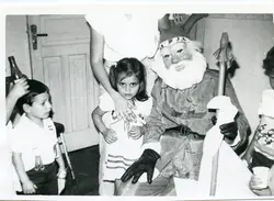 Fotografia em preto e branco capturando uma celebração natalina com uma criança posando ao lado de uma pessoa fantasiada de Papai Noel. O cenário sugere um ambiente doméstico ou escolar, típico de festividades de final de ano em meados do século XX.