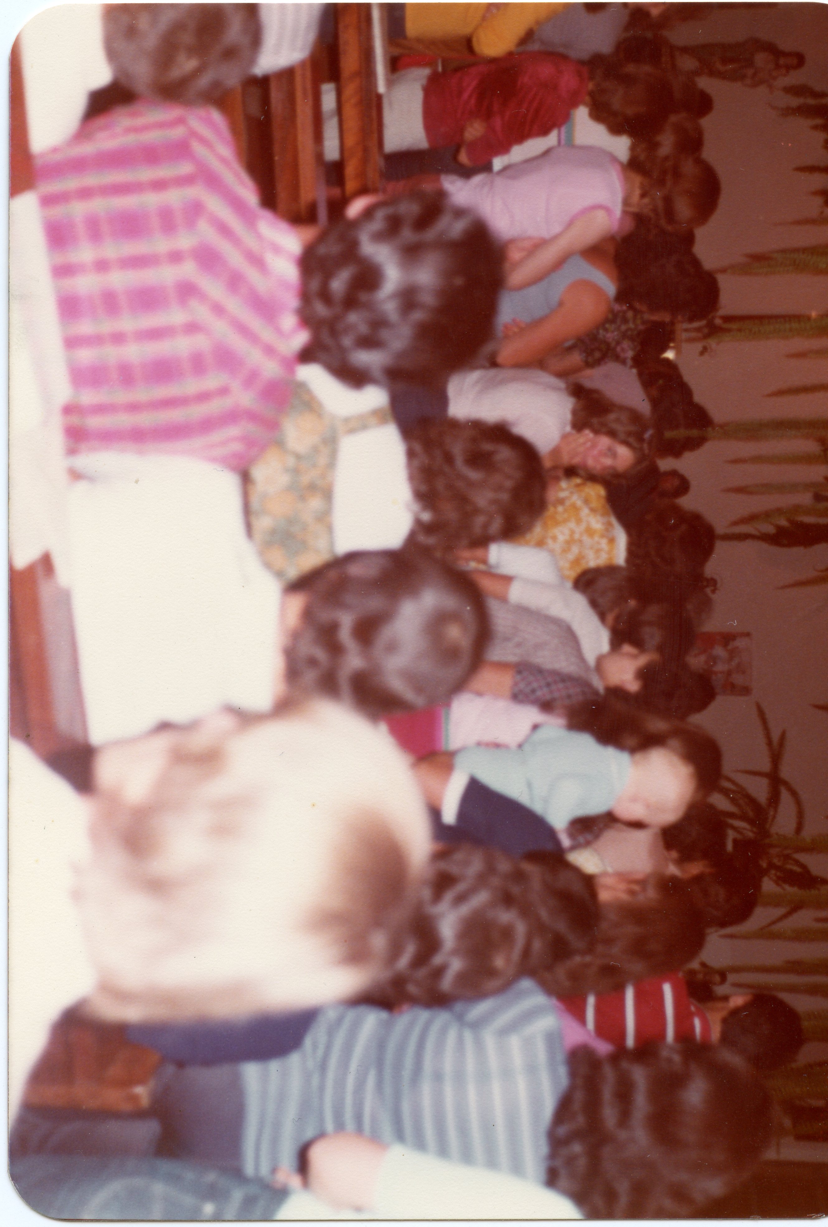 Uma fotografia colorida, possivelmente da década de 1980, que mostra um grupo de pessoas sentadas em bancos, sugerindo um ambiente de igreja ou reunião comunitária. A imagem está orientada verticalmente e apresenta um foco suave, com vestimentas típicas da época e uma atmosfera de encontro social ou religioso.