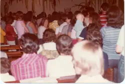 Uma fotografia colorida, possivelmente da década de 1980, que mostra um grupo de pessoas sentadas em bancos, sugerindo um ambiente de igreja ou reunião comunitária. A imagem está orientada verticalmente e apresenta um foco suave, com vestimentas típicas da época e uma atmosfera de encontro social ou religioso.