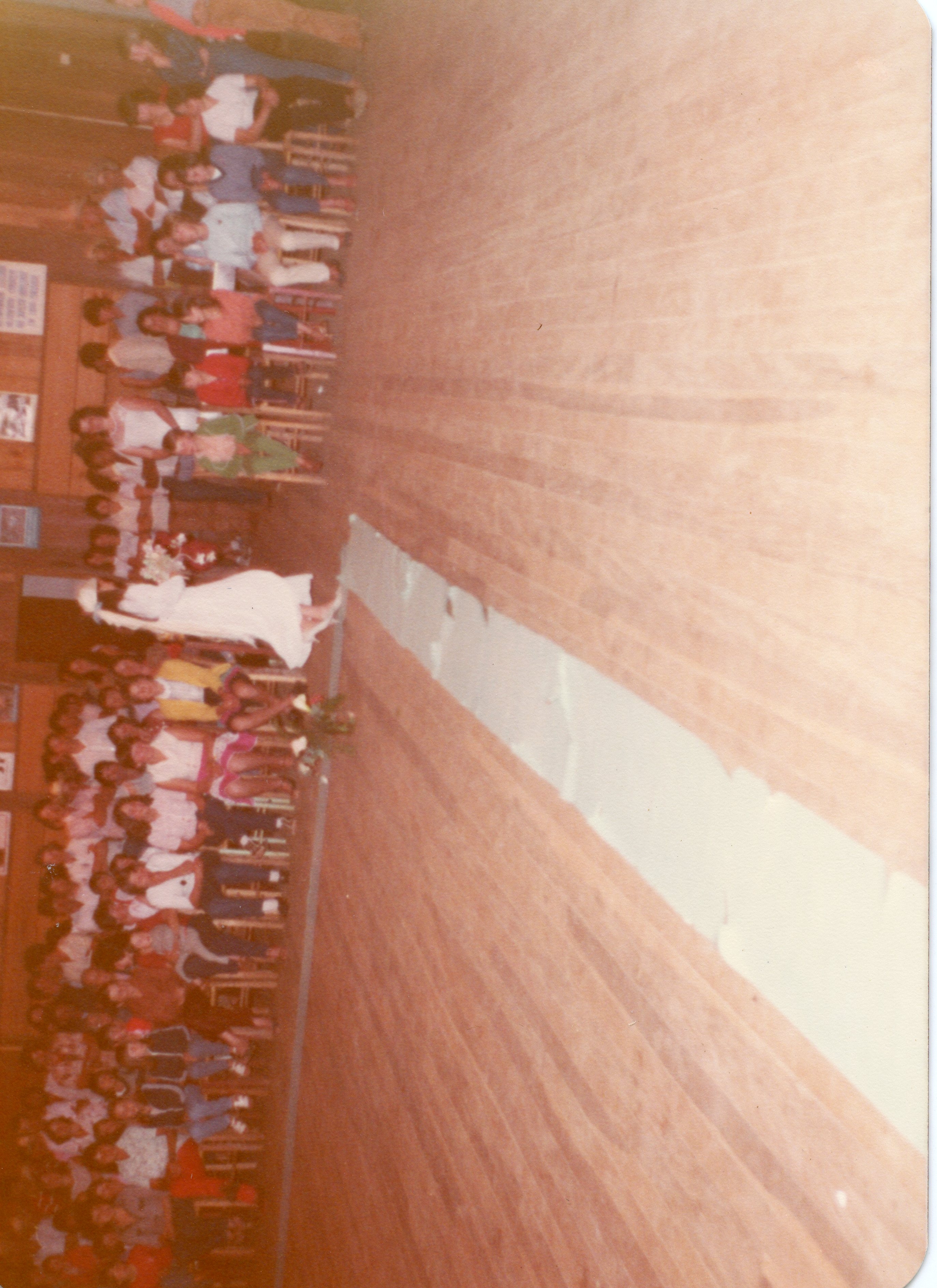 Fotografia colorida de época, possivelmente das décadas de 1970 ou 1980, capturando uma cerimônia de casamento em um salão com piso de madeira. Uma noiva caminha pelo corredor central enquanto convidados sentados em cadeiras de madeira observam a cena, refletindo um evento social típico de comunidades brasileiras.