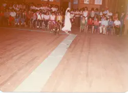 Fotografia colorida de época, possivelmente das décadas de 1970 ou 1980, capturando uma cerimônia de casamento em um salão com piso de madeira. Uma noiva caminha pelo corredor central enquanto convidados sentados em cadeiras de madeira observam a cena, refletindo um evento social típico de comunidades brasileiras.