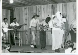 Fotografia em preto e branco retratando um grupo de pessoas em um ambiente interno com paredes de madeira, possivelmente um salão comunitário ou residência rural. O registro sugere uma reunião social ou celebração, com a presença de músicos tocando violão, indicando um contexto cultural típico de pequenas cidades brasileiras em meados do século XX.