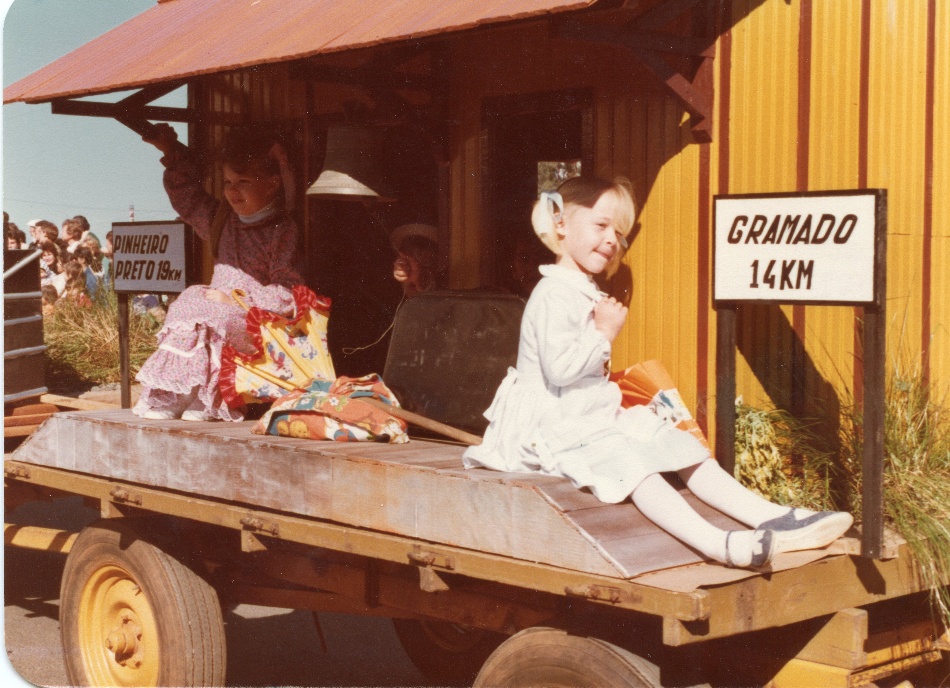 Uma fotografia colorida que mostra duas crianças vestidas com roupas de época, sentadas em uma estrutura de madeira que simula uma estação ferroviária ou carroça. O cenário inclui placas indicativas de distância para Gramado e Pinheiro Preto, sugerindo um evento cultural ou turístico, possivelmente na década de 1980.