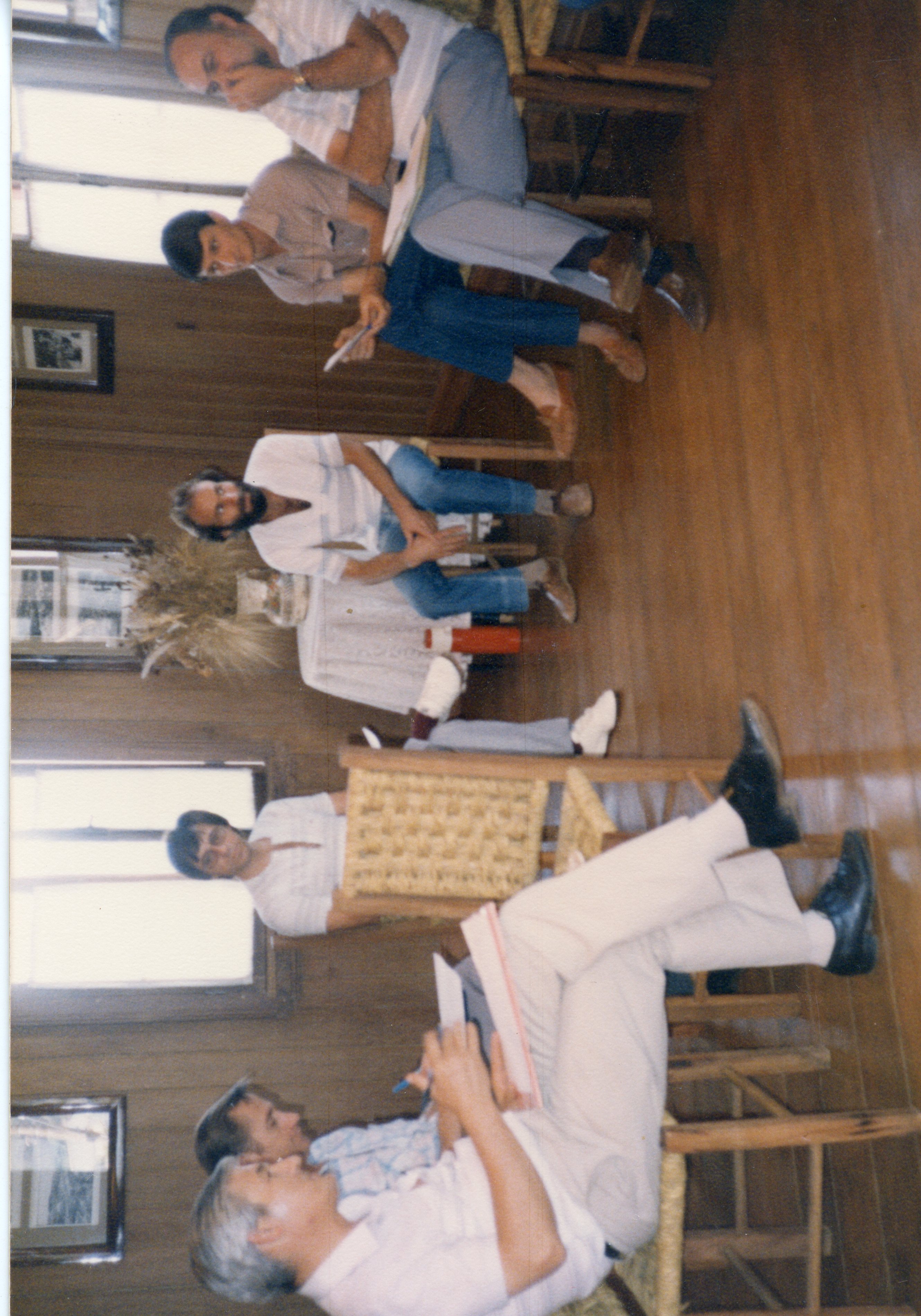 Uma fotografia de um grupo de homens sentados em uma sala com paredes de madeira, possivelmente durante uma reunião ou palestra informal. As vestimentas, incluindo camisas polo e calças jeans, sugerem um contexto brasileiro das décadas de 1980 ou 1990.