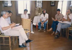 Uma fotografia de um grupo de homens sentados em uma sala com paredes de madeira, possivelmente durante uma reunião ou palestra informal. As vestimentas, incluindo camisas polo e calças jeans, sugerem um contexto brasileiro das décadas de 1980 ou 1990.