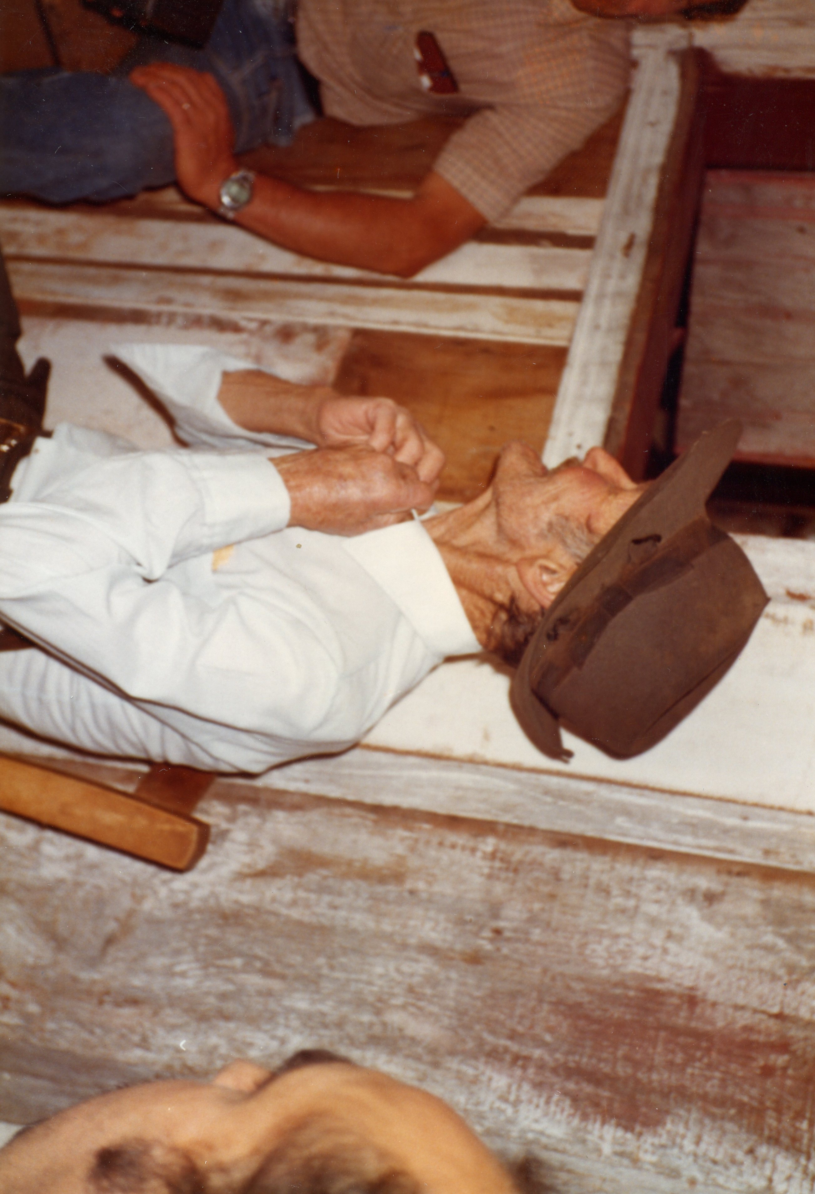 Uma fotografia em ângulo zenital mostrando um homem idoso deitado, vestindo camisa branca e chapéu, possivelmente em um ambiente rural ou de velório. A imagem apresenta características de fotografia amadora colorida das décadas de 1970 ou 1980, com a presença de outras pessoas ao redor.