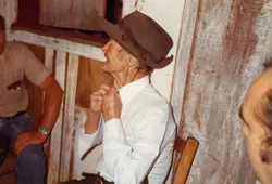Uma fotografia em ângulo zenital mostrando um homem idoso deitado, vestindo camisa branca e chapéu, possivelmente em um ambiente rural ou de velório. A imagem apresenta características de fotografia amadora colorida das décadas de 1970 ou 1980, com a presença de outras pessoas ao redor.