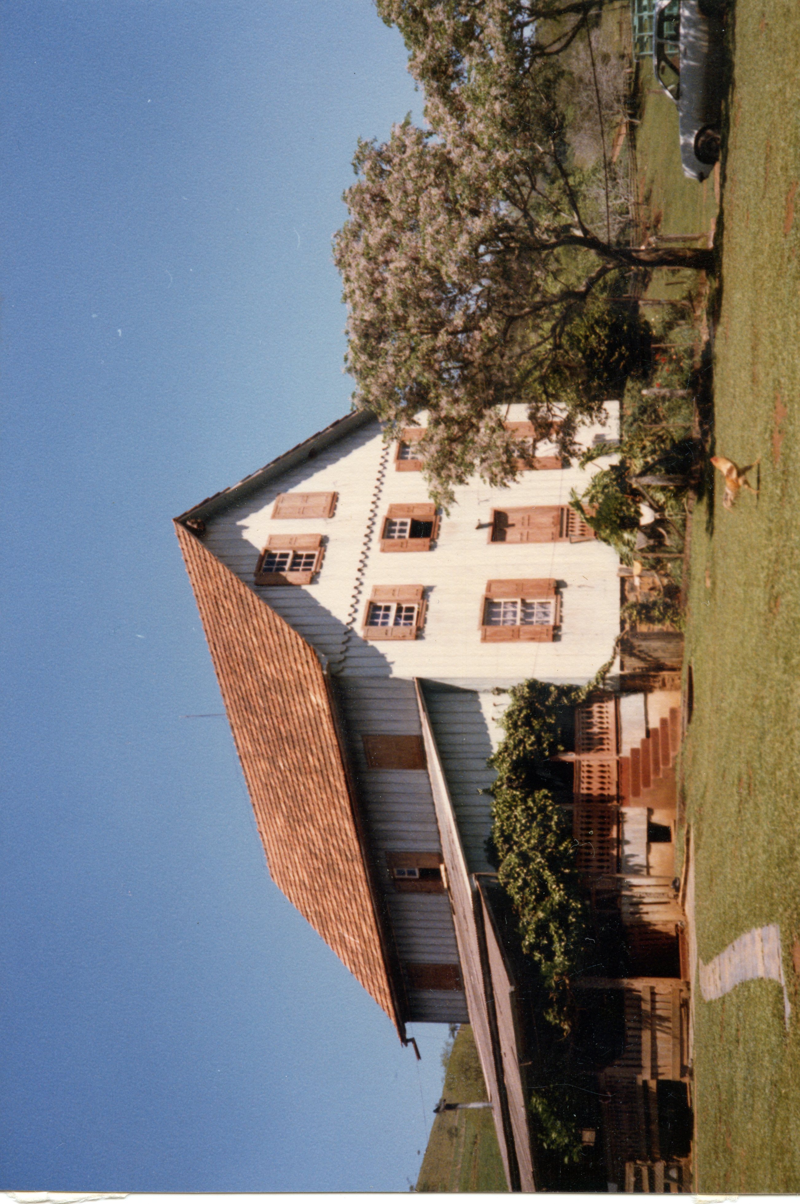 Uma fotografia colorida de uma casa de madeira de dois andares com telhado de telhas cerâmicas, típica da arquitetura rural de colonização europeia no sul do Brasil. A residência possui janelas com venezianas de madeira e está situada em um ambiente campestre, com uma árvore florida ao lado e um gramado bem cuidado.