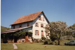 Uma fotografia colorida de uma casa de madeira de dois andares com telhado de telhas cerâmicas, típica da arquitetura rural de colonização europeia no sul do Brasil. A residência possui janelas com venezianas de madeira e está situada em um ambiente campestre, com uma árvore florida ao lado e um gramado bem cuidado.