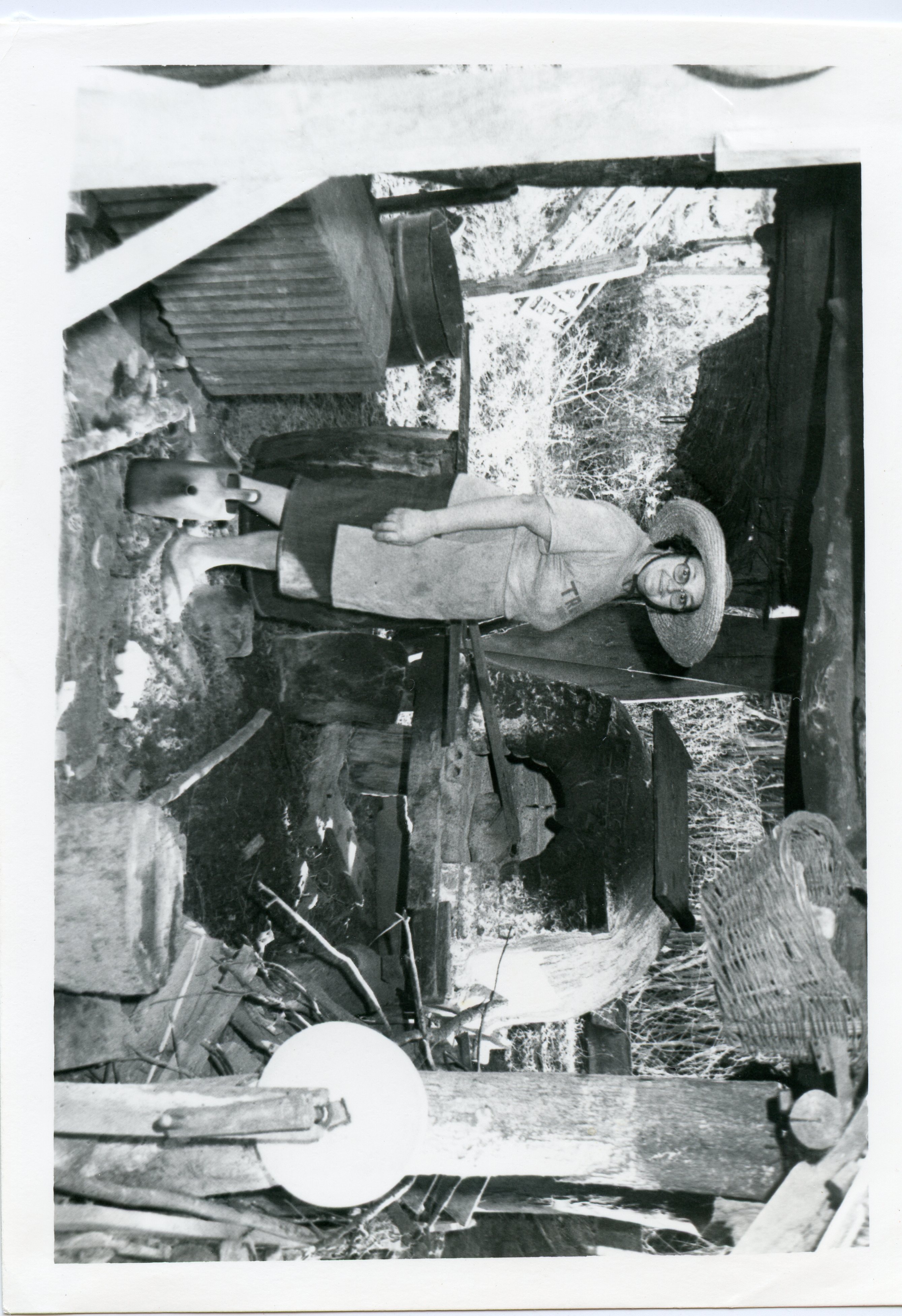 Uma fotografia em preto e branco que retrata uma mulher usando chapéu de palha e óculos, posicionada em um ambiente rústico que parece ser um forno de barro ou área de trabalho rural. A cena sugere atividades domésticas ou artesanais típicas da vida no campo, possivelmente em meados do século XX.