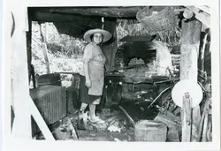 Uma fotografia em preto e branco que retrata uma mulher usando chapéu de palha e óculos, posicionada em um ambiente rústico que parece ser um forno de barro ou área de trabalho rural. A cena sugere atividades domésticas ou artesanais típicas da vida no campo, possivelmente em meados do século XX.
