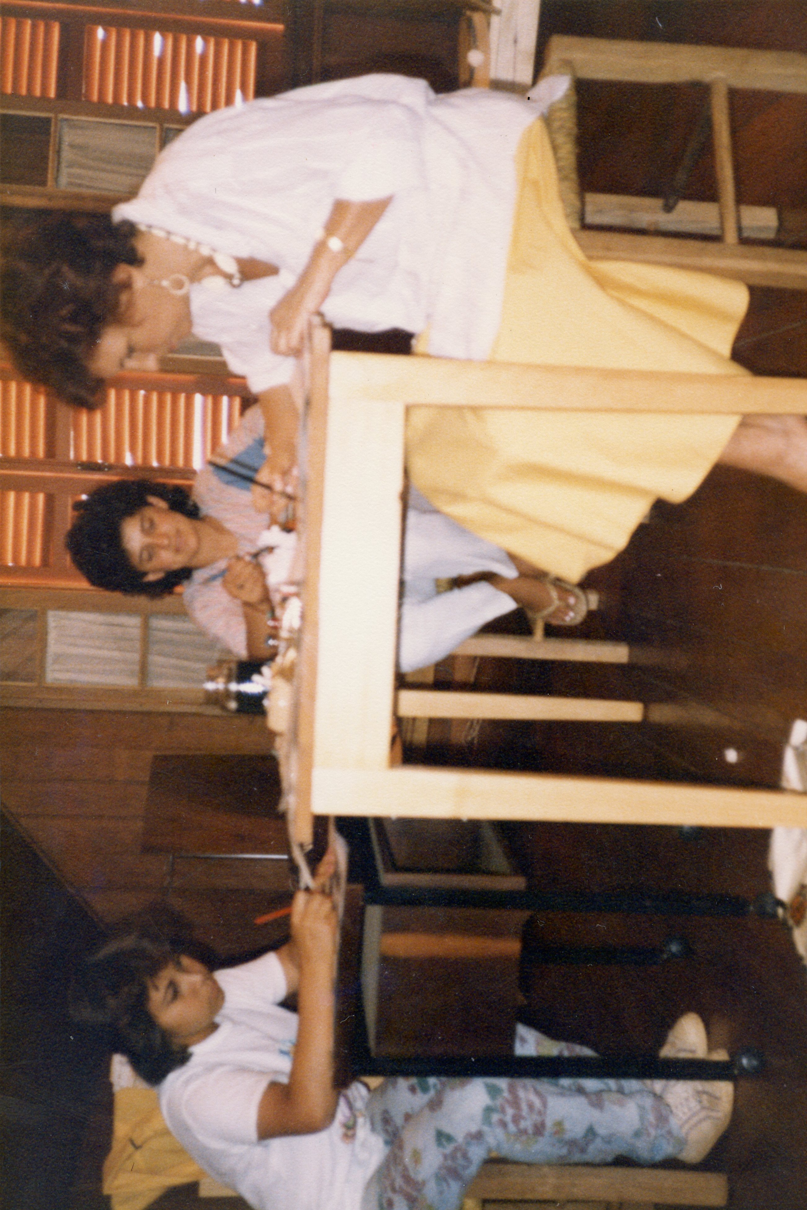 Três mulheres estão reunidas em torno de uma mesa de madeira, possivelmente em uma atividade de artesanato ou trabalho manual. A iluminação sugere um ambiente interno, talvez uma casa ou oficina. As vestimentas e o estilo da fotografia indicam que a imagem pode ter sido tirada nas décadas de 1970 ou 1980.
