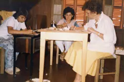 Três mulheres estão reunidas em torno de uma mesa de madeira, possivelmente em uma atividade de artesanato ou trabalho manual. A iluminação sugere um ambiente interno, talvez uma casa ou oficina. As vestimentas e o estilo da fotografia indicam que a imagem pode ter sido tirada nas décadas de 1970 ou 1980.