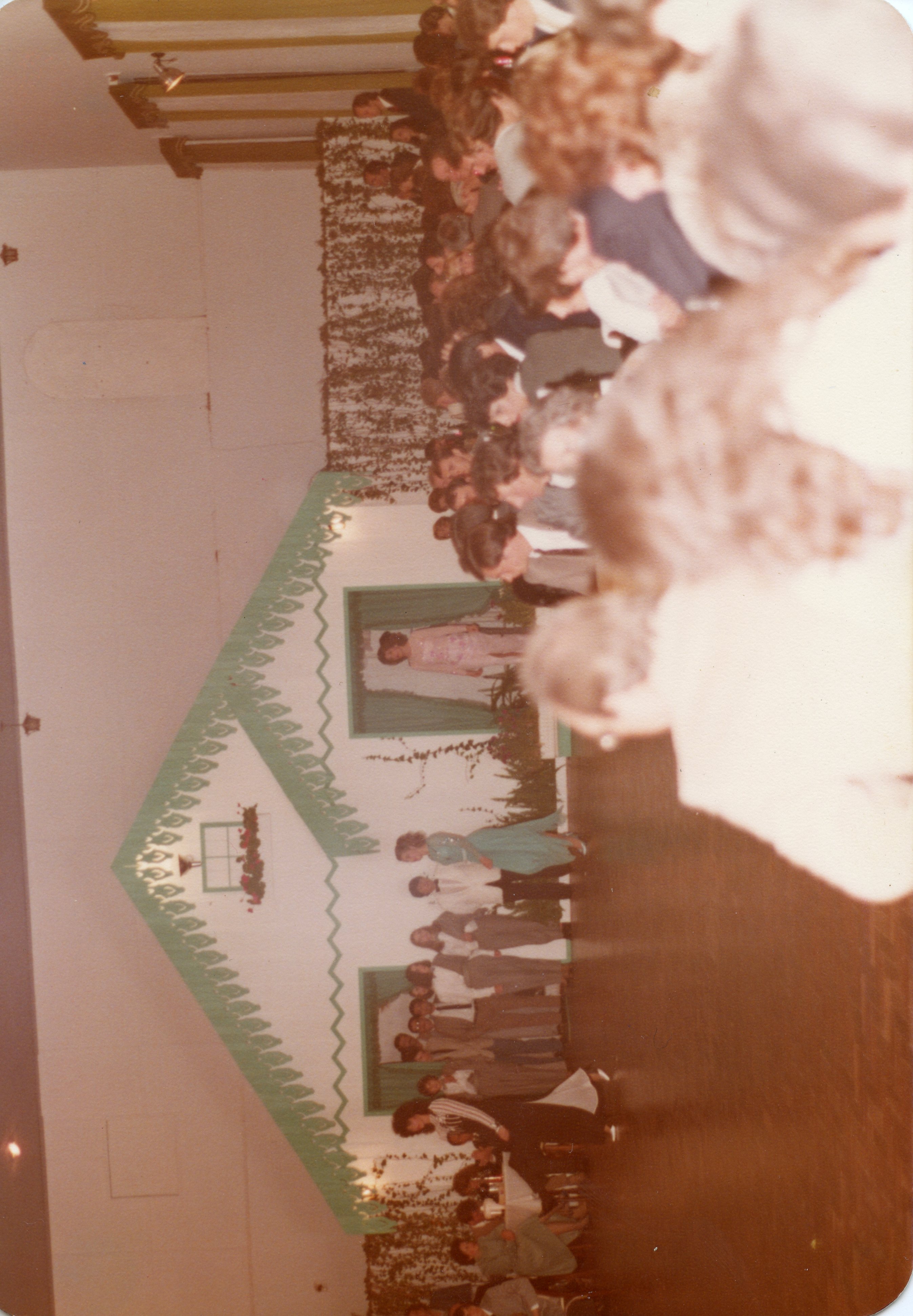 A fotografia mostra um grupo de pessoas reunidas em um evento, possivelmente uma festa ou celebração, com uma decoração que remete a uma casa com telhado verde e detalhes em folhagem. A vestimenta das pessoas sugere que a foto foi tirada nas décadas de 1970 ou 1980. Há uma figura em destaque em um quadro, possivelmente um homenageado ou uma representação simbólica.