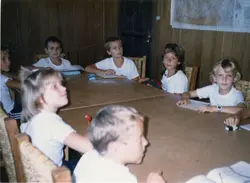 Uma fotografia colorida de uma sala de aula com crianças sentadas ao redor de uma grande mesa de madeira. O ambiente possui paredes revestidas de madeira e um mapa na parede, sugerindo uma escola ou centro comunitário, provavelmente na década de 1990.
