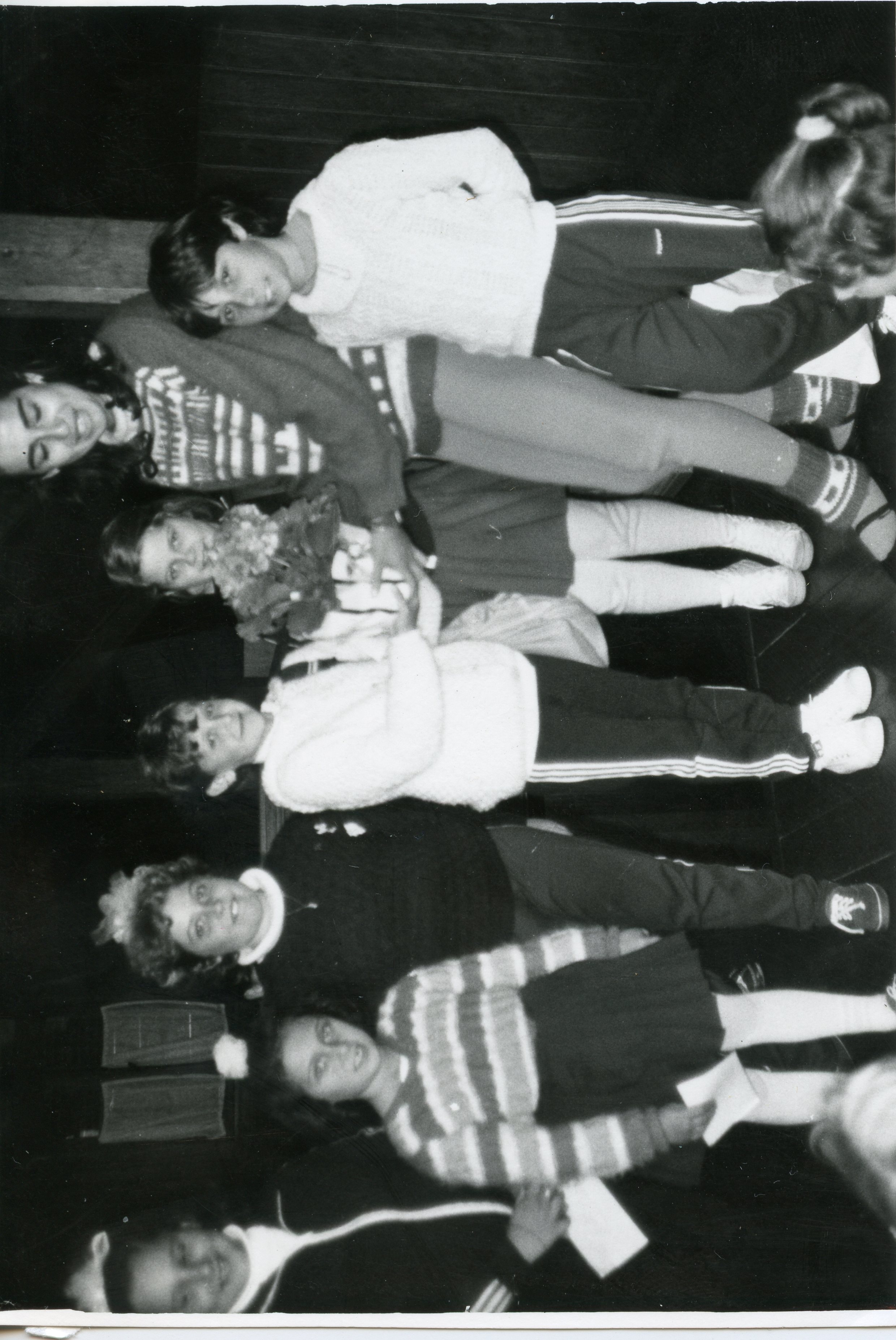 Fotografia em preto e branco retratando um grupo de crianças e jovens em um ambiente interno, possivelmente uma escola ou centro comunitário. As vestimentas, incluindo agasalhos de lã e calças esportivas, sugerem um contexto escolar brasileiro entre as décadas de 1970 e 1980.