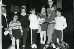 Fotografia em preto e branco retratando um grupo de crianças e jovens em um ambiente interno, possivelmente uma escola ou centro comunitário. As vestimentas, incluindo agasalhos de lã e calças esportivas, sugerem um contexto escolar brasileiro entre as décadas de 1970 e 1980.