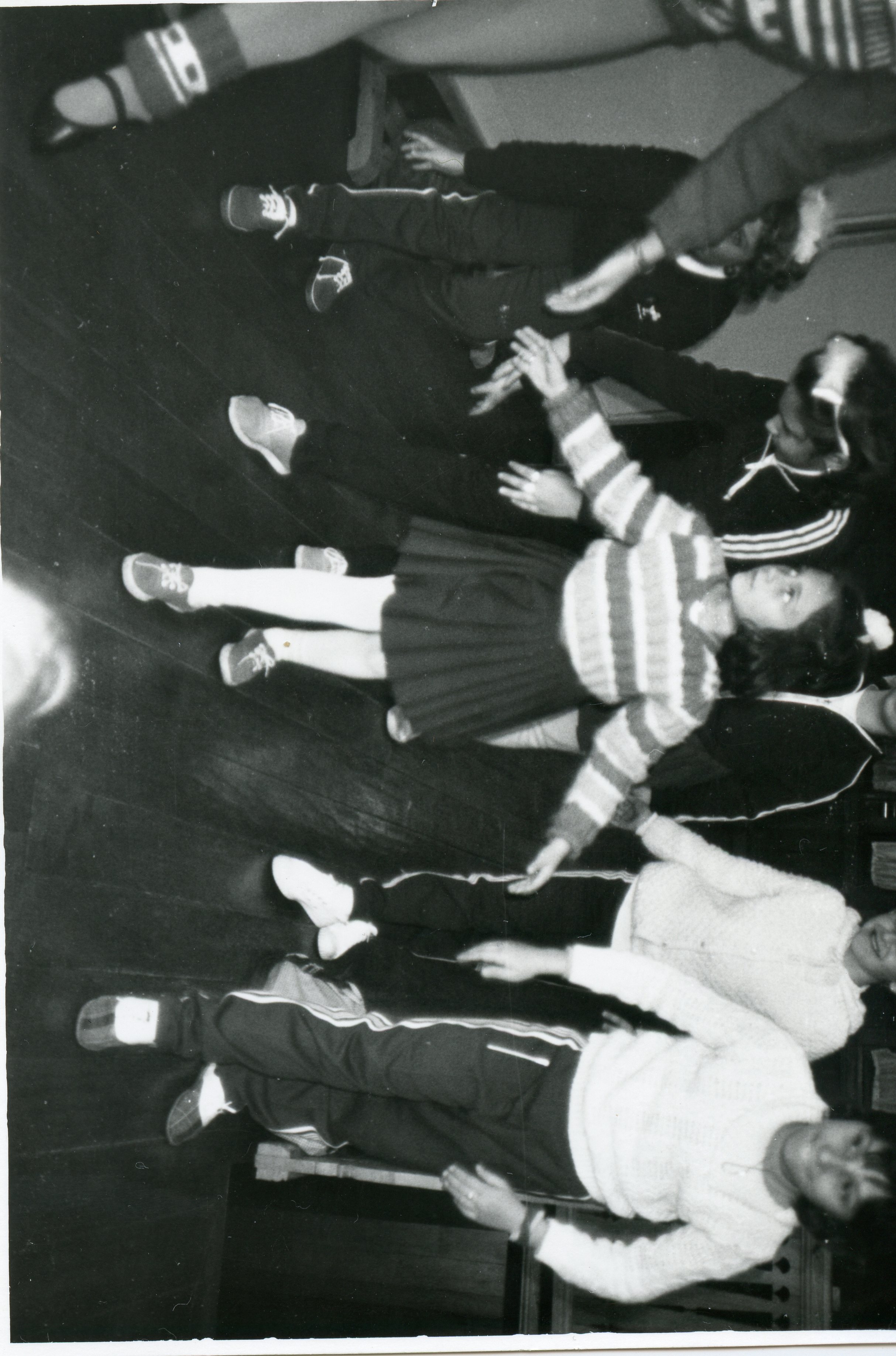 Fotografia em preto e branco capturada de um ângulo superior, mostrando um grupo de jovens ou crianças em um ambiente interno com piso de madeira, possivelmente participando de uma atividade escolar ou recreativa. As vestimentas, incluindo agasalhos de lã e calças esportivas, sugerem um contexto brasileiro entre as décadas de 1970 e 1980.