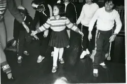 Fotografia em preto e branco capturada de um ângulo superior, mostrando um grupo de jovens ou crianças em um ambiente interno com piso de madeira, possivelmente participando de uma atividade escolar ou recreativa. As vestimentas, incluindo agasalhos de lã e calças esportivas, sugerem um contexto brasileiro entre as décadas de 1970 e 1980.