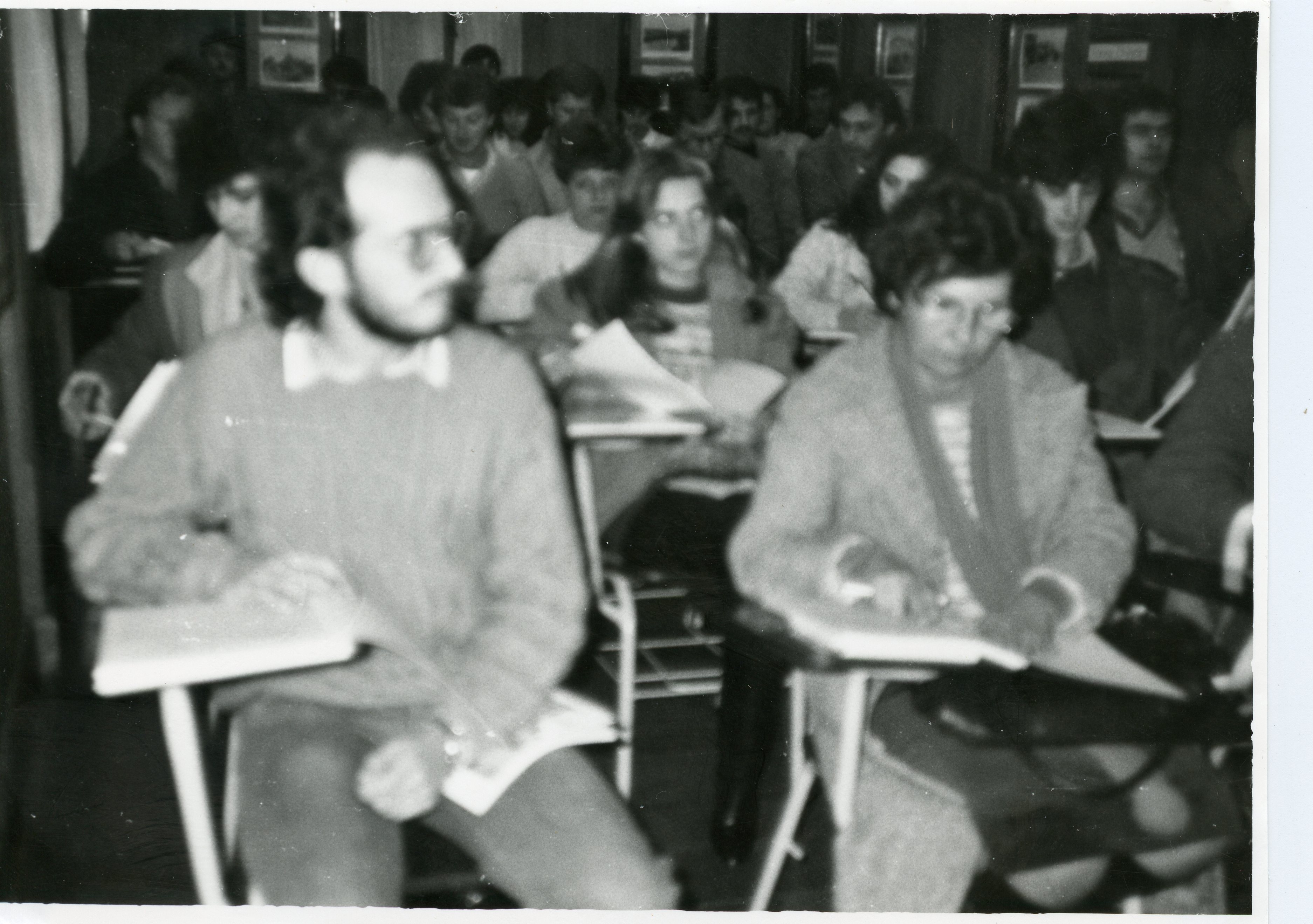 Uma fotografia em preto e branco que retrata uma sala de aula ou auditório lotado, com estudantes sentados em carteiras escolares individuais. O estilo das vestimentas, como suéteres e cortes de cabelo, sugere um contexto acadêmico brasileiro provavelmente entre as décadas de 1970 e 1980.