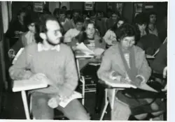 Uma fotografia em preto e branco que retrata uma sala de aula ou auditório lotado, com estudantes sentados em carteiras escolares individuais. O estilo das vestimentas, como suéteres e cortes de cabelo, sugere um contexto acadêmico brasileiro provavelmente entre as décadas de 1970 e 1980.