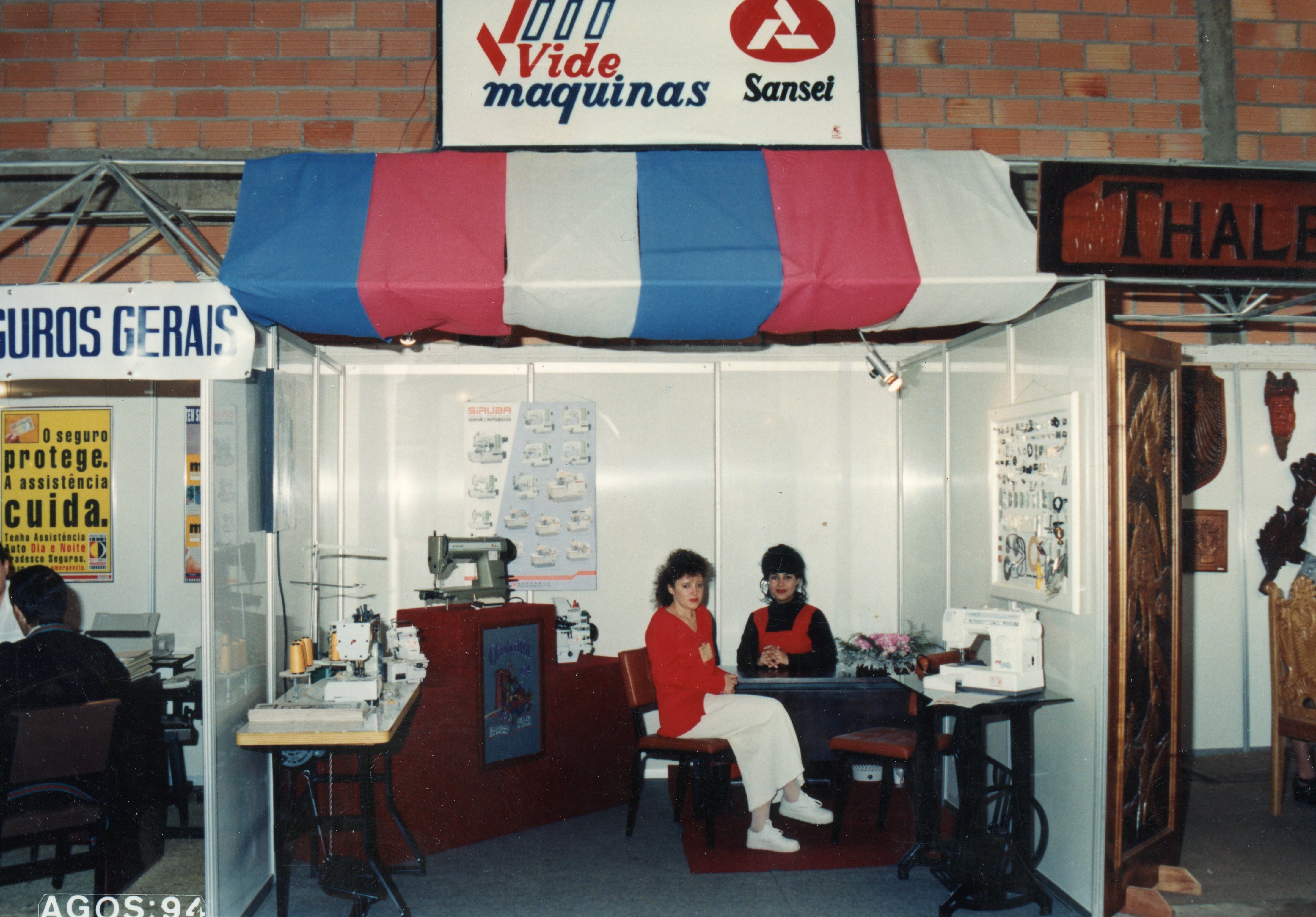 Uma fotografia de um estande comercial em uma feira, provavelmente em Videira, exibindo máquinas de costura da marca Sansei. Duas mulheres estão sentadas no estande, que apresenta decoração com toldo tricolor e paredes de divisórias modulares, datado de agosto de 1994.