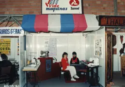 Uma fotografia de um estande comercial em uma feira, provavelmente em Videira, exibindo máquinas de costura da marca Sansei. Duas mulheres estão sentadas no estande, que apresenta decoração com toldo tricolor e paredes de divisórias modulares, datado de agosto de 1994.