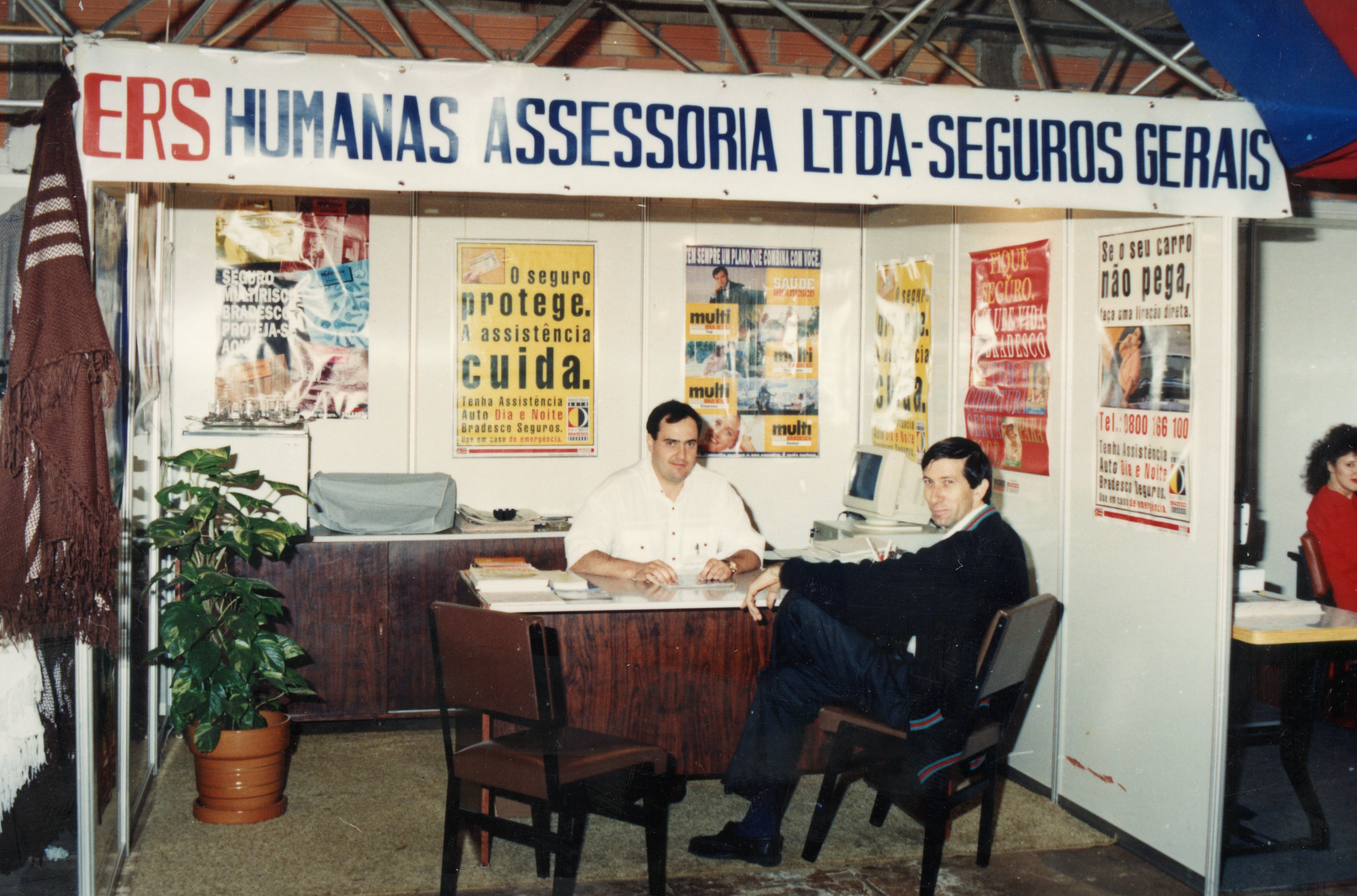 Uma fotografia de um estande de feira de negócios ou exposição comercial, provavelmente da década de 1990, apresentando a empresa 'Humanas Assessoria Ltda - Seguros Gerais'. Dois homens estão sentados atrás de uma mesa de atendimento, cercados por cartazes promocionais de seguros e equipamentos de escritório da época, como um monitor de computador antigo.