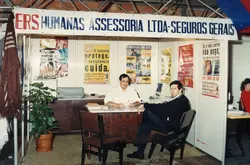 Uma fotografia de um estande de feira de negócios ou exposição comercial, provavelmente da década de 1990, apresentando a empresa 'Humanas Assessoria Ltda - Seguros Gerais'. Dois homens estão sentados atrás de uma mesa de atendimento, cercados por cartazes promocionais de seguros e equipamentos de escritório da época, como um monitor de computador antigo.