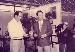 Uma fotografia em tons sépia retratando um evento promocional ou feira, possivelmente em um ambiente comercial ou industrial. Dois homens adultos conversam enquanto um jovem segura uma câmera de vídeo profissional, sugerindo uma cobertura jornalística ou registro de evento na década de 1980 ou início de 1990.