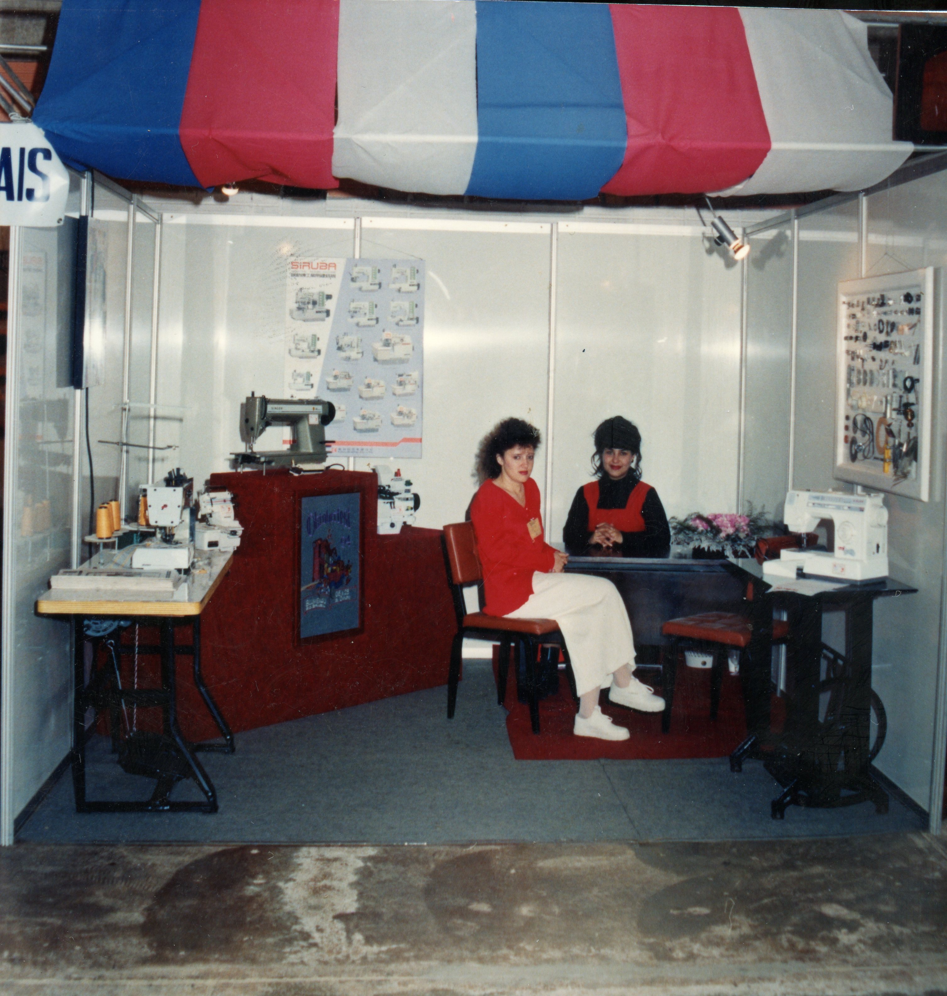 Uma fotografia colorida de um estande de feira comercial focado em máquinas de costura, provavelmente da década de 1990. Duas mulheres estão sentadas no estande, que apresenta cartazes informativos, mostruários de peças e diversas máquinas de costura expostas sob um toldo tricolor.