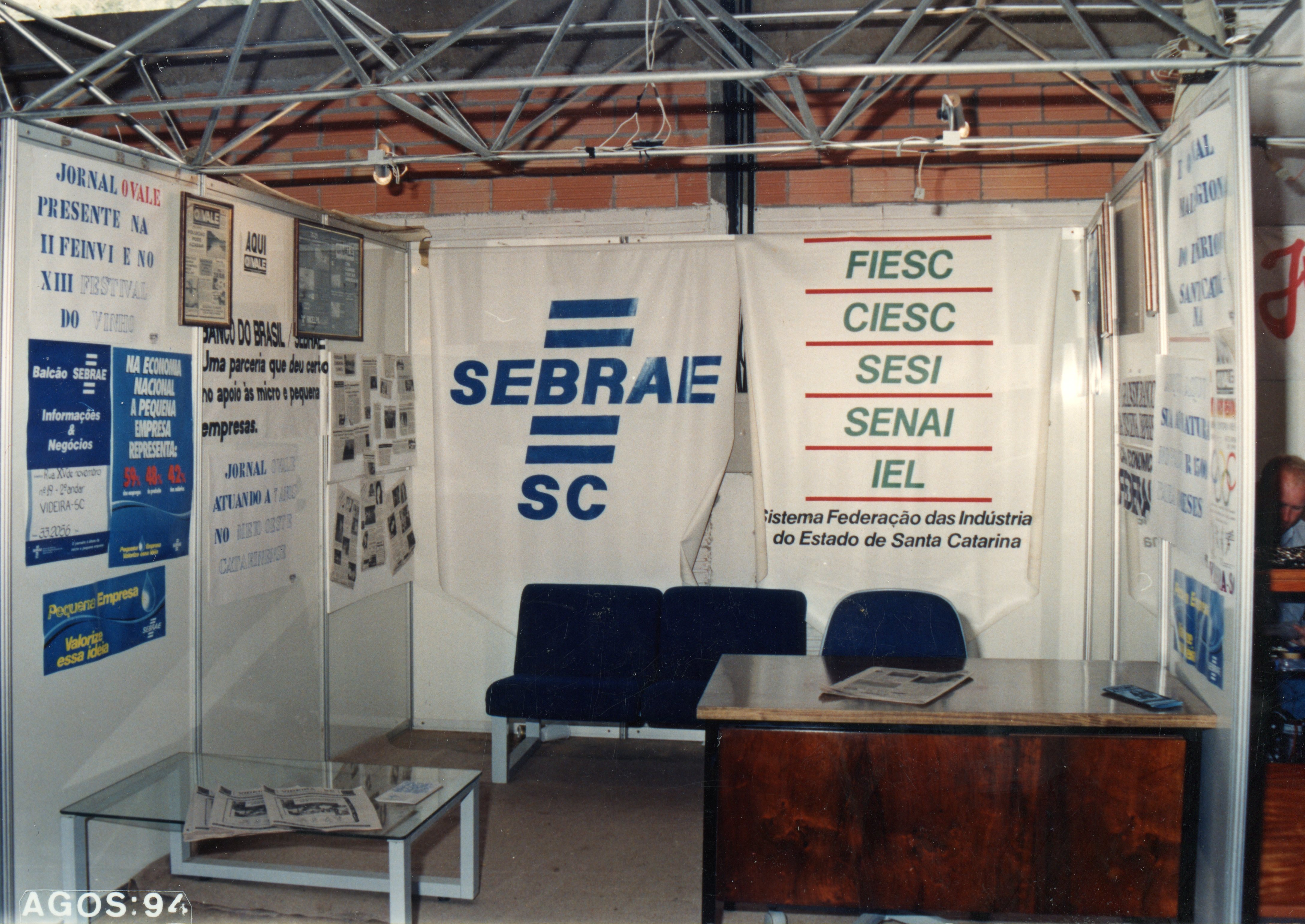 Uma fotografia de um estande institucional em uma feira de negócios, provavelmente a FEINVI em Videira, Santa Catarina. O espaço exibe logotipos de entidades como SEBRAE e FIESC, com cartazes informativos e mobiliário de escritório, datado de agosto de 1994.