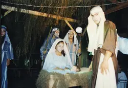 Uma encenação teatral de um presépio vivo, provavelmente realizada em um contexto comunitário ou escolar. Os participantes vestem trajes que remetem à época bíblica, com uma jovem representando Maria ao lado de um bebê em uma manjedoura, sob uma estrutura rústica de palha.