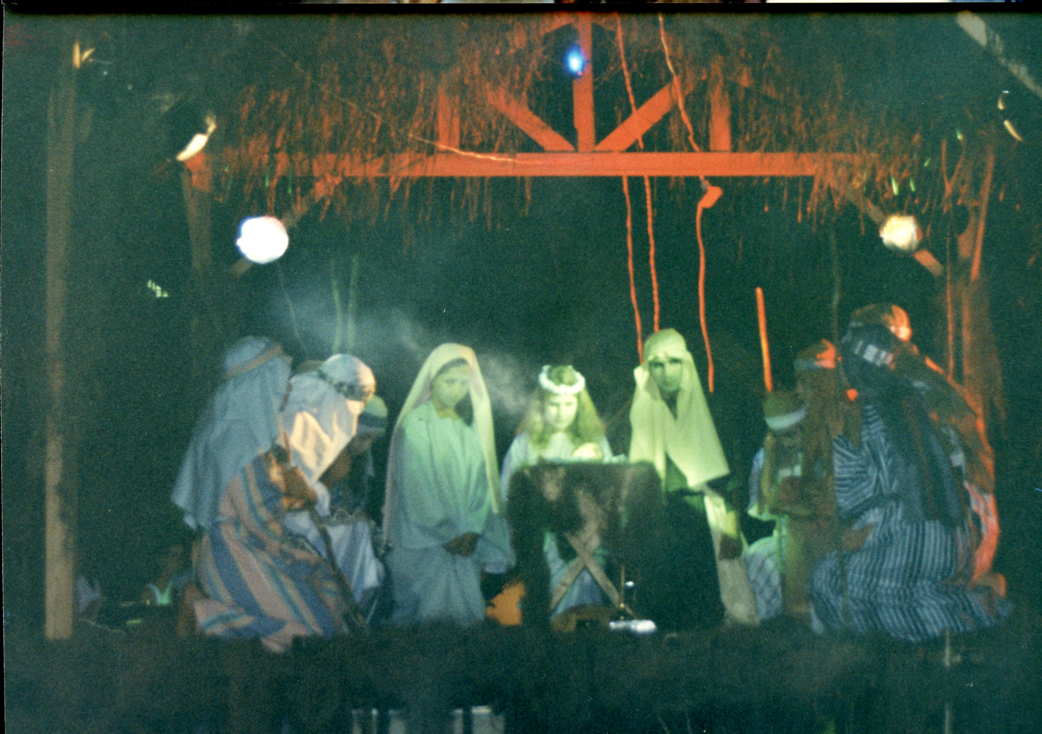 Uma fotografia que retrata uma encenação teatral de um presépio vivo, provavelmente realizada em um contexto comunitário ou escolar. Os participantes vestem trajes típicos que remetem à época bíblica, sob uma estrutura rústica de madeira e palha, iluminada artificialmente em um ambiente noturno.