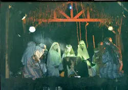 Uma fotografia que retrata uma encenação teatral de um presépio vivo, provavelmente realizada em um contexto comunitário ou escolar. Os participantes vestem trajes típicos que remetem à época bíblica, sob uma estrutura rústica de madeira e palha, iluminada artificialmente em um ambiente noturno.