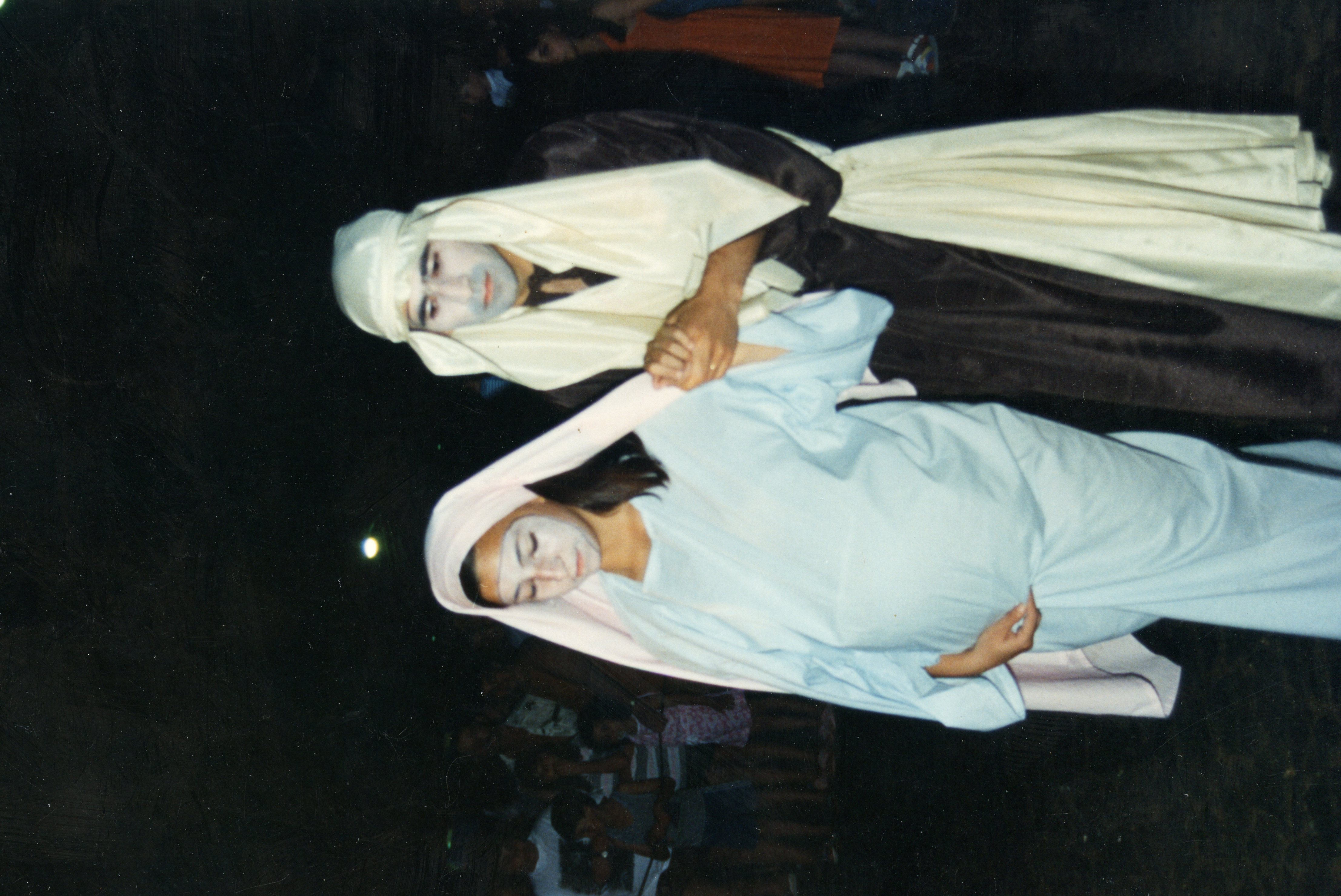 Uma fotografia que retrata uma encenação teatral, provavelmente uma representação bíblica ou presépio vivo, com dois atores caracterizados como José e Maria. Os atores utilizam vestimentas típicas de época e maquiagem teatral, em um ambiente noturno que sugere uma celebração comunitária ou escolar.