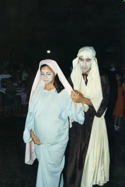 Uma fotografia que retrata uma encenação teatral, provavelmente uma representação bíblica ou presépio vivo, com dois atores caracterizados como José e Maria. Os atores utilizam vestimentas típicas de época e maquiagem teatral, em um ambiente noturno que sugere uma celebração comunitária ou escolar.