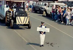 Uma cena de desfile cívico ou festividade urbana, onde um jovem uniformizado carrega uma placa indicando 'Trem Lions Centro'. Ao fundo, observa-se um veículo adaptado como trem e uma multidão de espectadores, sugerindo um evento comunitário típico de cidades brasileiras nas décadas de 1980 ou 1990.