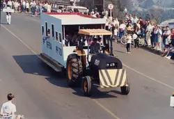 Uma fotografia colorida registrando um desfile ou evento público, possivelmente em uma cidade brasileira, onde um trator adaptado puxa um vagão decorado com a inscrição 'RFFSA PP 720'. O veículo faz alusão a uma locomotiva ferroviária, com pessoas sentadas dentro do vagão e uma multidão observando nas margens da via, datada de agosto de 1994.