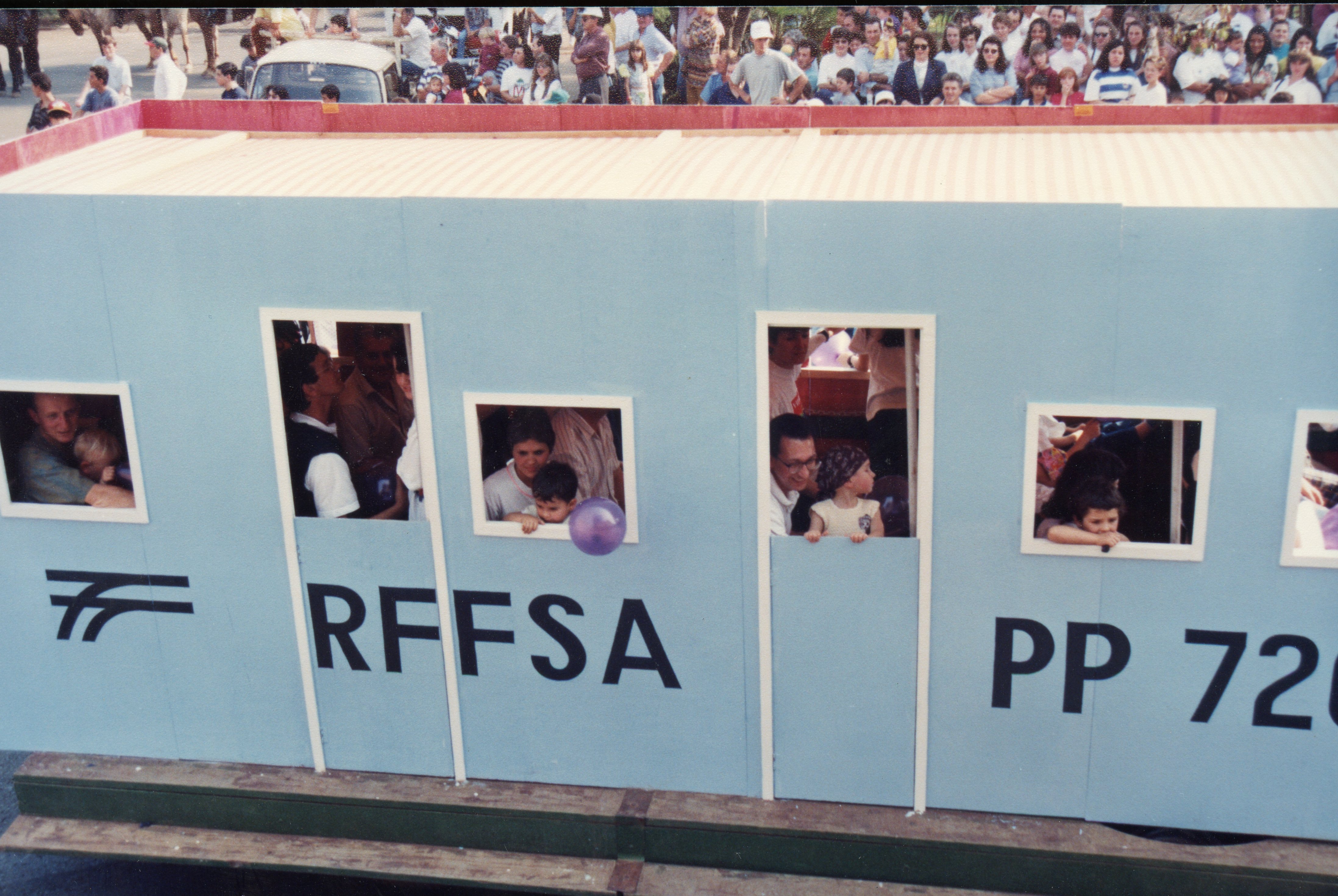 Uma fotografia colorida que registra um desfile ou evento público, onde um carro alegórico decorado como um vagão de trem da RFFSA (Rede Ferroviária Federal S.A.) transporta diversas pessoas, incluindo crianças. O cenário sugere uma celebração comunitária, possivelmente em uma cidade brasileira com tradição ferroviária, datada provavelmente da década de 1990 pelas vestimentas e qualidade da imagem.